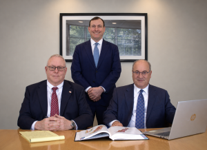 Three men in business suits pose in a conference room; one stands behind two seated at a table with a magazine and an open laptop visible on the right side.