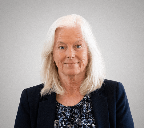 Portrait of an older woman with white hair wearing a navy blazer and floral blouse against a gray backdrop, looking at the camera with a gentle smile.