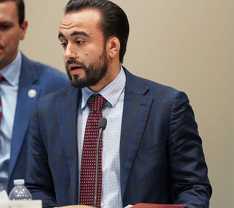 Gerard M. Damiani III Man in a navy blue checkered suit speaks into a small microphone during a panel discussion, seated at a table with a water bottle nearby.